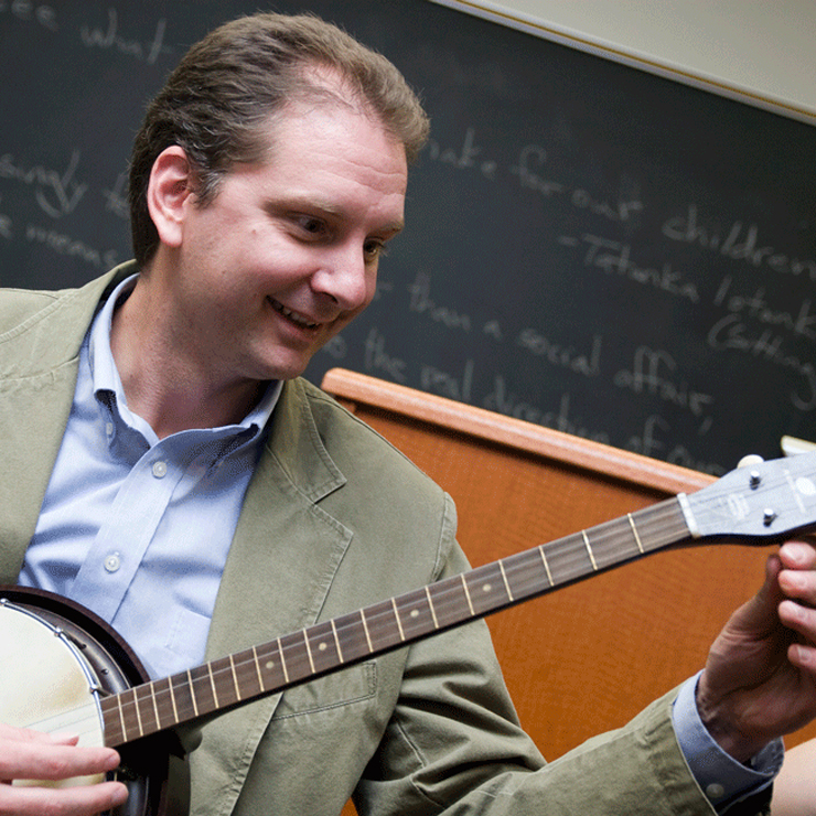 A white man with short hair and wearing a tan suit sits in a classroom tuning a banjo