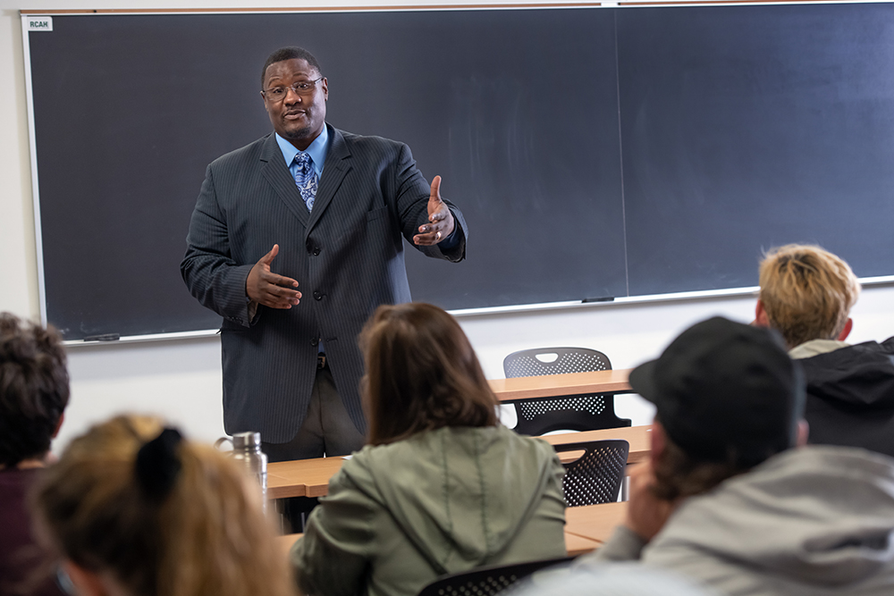 Professor Kevin Brooks, an african american professor, stands in front of his class in a suit and shares information with students.