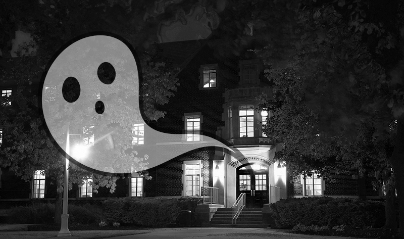 Image shows a brick building, Phillips Hall, at night lit up with every light on. A cartoonish ghost escapes from the doorway into the night.