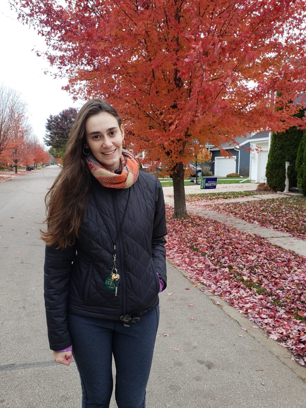 A young White Latina woman standing in the middle of a neighborhood street near a tree with orange fall foliage, wearing a black jacket, blue pants, and an orange scarf.